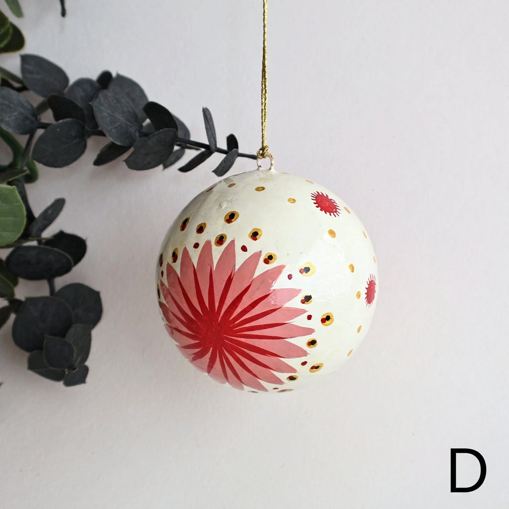 A spherical Christmas ornament with a cream background, featuring a large red starburst design, small red flowers, and gold polka dots, hanging from a gold string against a white wall with eucalyptus leaves on the left.