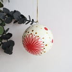 A spherical Christmas ornament with a cream background, featuring a large red starburst design, small red flowers, and gold polka dots, hanging from a gold string against a white wall with eucalyptus leaves on the left.